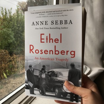 A close-up photo of a hand holding up Anne Sebba's biography (paperback edition), "Ethel Rosenberg: An American Tragedy" against the backdrop of a window, beige curtains & a white windowsill. Out the window, greenery & gray skies are visible. The cover of the book depicts a black & white photo of Ethel stepping out of a car, with three men in suits around her, looking towards her from outside the car.