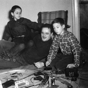 Black and white photo from 1954 of Abel Meeropol (middle) playing with his two sons, Robert (left) and Michael (right) in front of a model train set. 