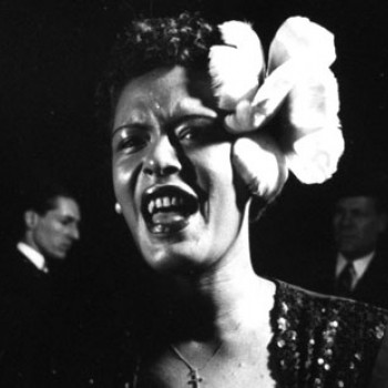 Black and white photo of Billie Holiday singing at the microphone, close-up, cropped to see only her face. Her hair is done up with her signature white gardenia flower tucked behind her ear.