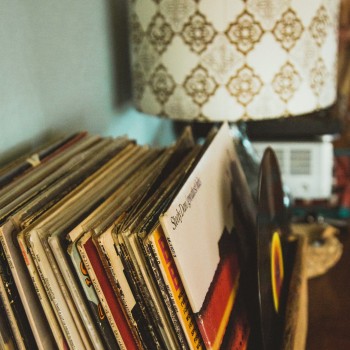 A stack of vinyl albums in their sleeves face the right side of the photo. The last album in the stack is out of its sleeve. They are organized on a tabletop in front of a lamp with a white a gold patterned lampshade, all inside a pale blue painted room. 
