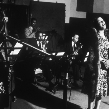 Black and white photo or Billie Holiday singing on stage in front of a microphone. She wears a floral patterned dress and a fur stole. Her eyes are closed, head tilted to the left, she is singing. 4 band members accompany her including 2 saxophonists, 1 trumpet, & 1 upright bass player. All wear suits. A piano and music stands are visible.