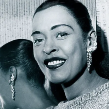 Close-up portrait of Billie Holiday shown looking away from a mirror behind her. She wears her long hair in a low ponytail, intricate jeweled earrings that hang low and crawl up the ear. She smiles. Her reflection is visible looking in the opposite direction.