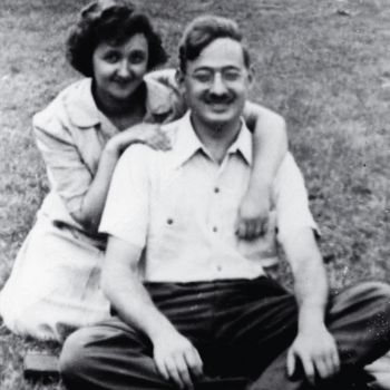 Black and white photo of Ethel and Julius Rosenberg seated outdoors. Ethel wears a light colored dress and kneels with her arms around Julius's shoulders. Julius sits cross-legged and wears a white shirt, dark pants, and glasses. They are both smiling.