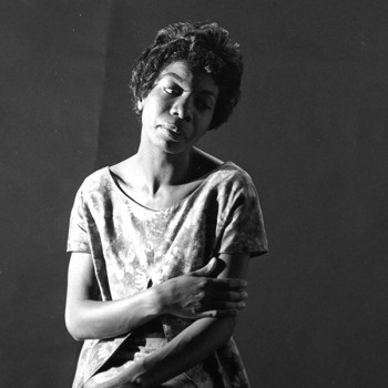 Portrait of Nina Simone in black and white, she is centered against a black backdrop. Her right arm crosses her body. She looks down and to her left.