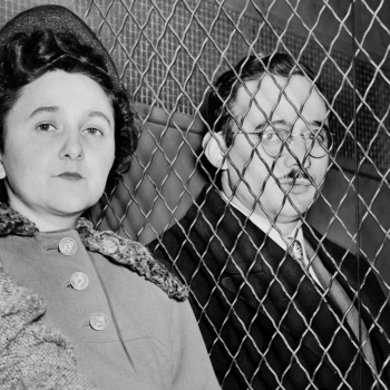 Close-up, black and white photo of Ethel & Julius Rosenberg at Sing Sing, seated and separated by a chain link fence, facing forward.