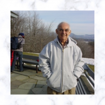 Photo of David Alman, cropped. He wears a zip up, gray hoodie. In the background is fencing, forest and mountain views.