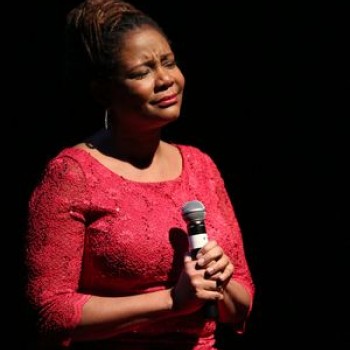 Tonya Pinkins, wearing a red dress with her hair done up in a bun, sits on stage with lights darkened to black behind her, spotlight on her. She faces slightly to the right and grips a microphone to her chest. Her eyes are closed, and her face is emotional.
