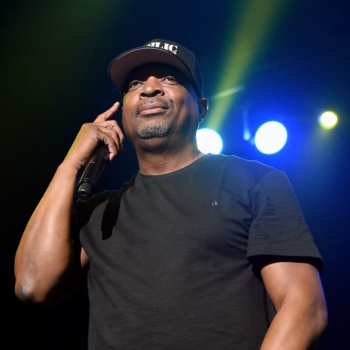 A photo of rapper Chuck D of Public Enemy on stage, wearing a black t-shirt and a black cap. He holds a microphone in his right hand close to his face. Stage lights flash behind him against a black backdrop.