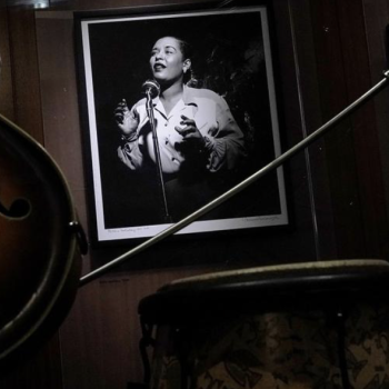 A framed, black and white photo of Billie Holiday hangs on a dark wall alongside musical memorabilia: a blues guitar, a saxophone, and a drum.