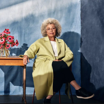 Angela Davis poses, seated beside a table with a vase of flowers.