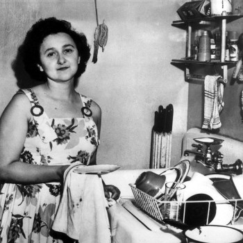 Ethel Rosenberg smiles as she stands next to a kitchen sink, with a plate and washcloth in hand, clean dishes piled in the drying rack beside her. She wears a sundress with a large, floral pattern.