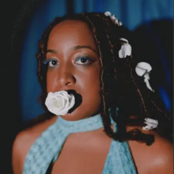 Singer Amahla in her music video "Enough" wearing a sleeveless aqua blue dress, white gardenia flower petals in her hair and a white rose in her mouth